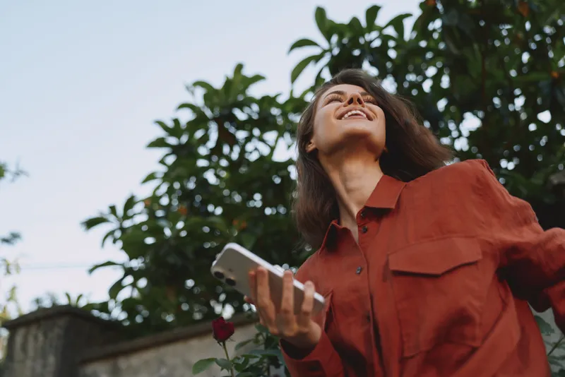 Young woman enjoying outdoor lifestyle, smiling up at the sky, holding a smartphone, wearing a red shirt, in a sunny garden setting, carefree and relaxed