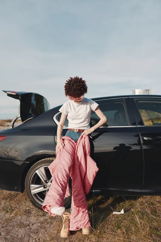 Outdoor scene with a person by car in casual fashion and street style, back view on a sunny day, showing summer vibe and a relaxed pose beside a black sedan with open trunk