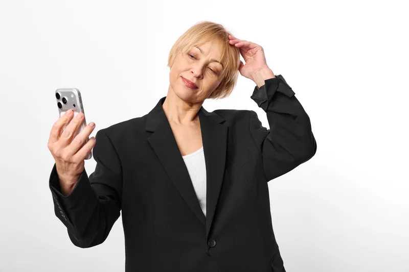 Woman businesswoman holding smartphone during phone call, hand at ear with gentle smile, studio portrait in black suit, clean white background, communication technology concept