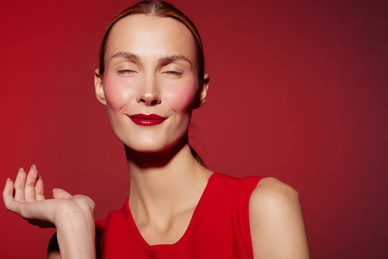 Smiling woman with closed eyes wearing red dress and lipstick poses against red background. Concept of beauty, fashion, confidence, and elegance in studio portrait.