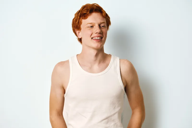 Young man with red hair in white tank top smiling and looking away. Casual male portrait isolated on plain white background with relaxed and confident pose.