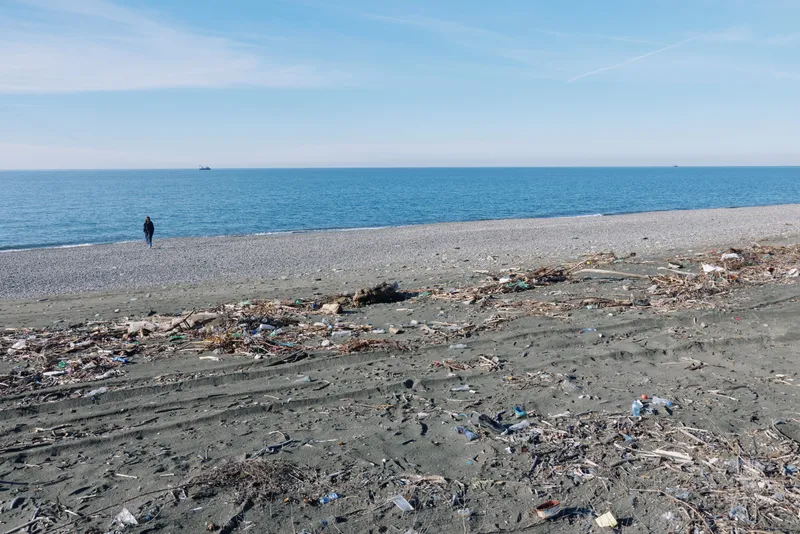 Beach, litter and trash litter the shoreline as waves meet the coast, with a solitary figure near the water under a clear sky, highlighting pollution and environmental awareness.