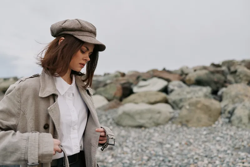 Young woman in vintage clothing and cap stands on rocky beach looking down. Overcast sky and casual autumn outfit create a calm, reflective mood in natural outdoor setting.