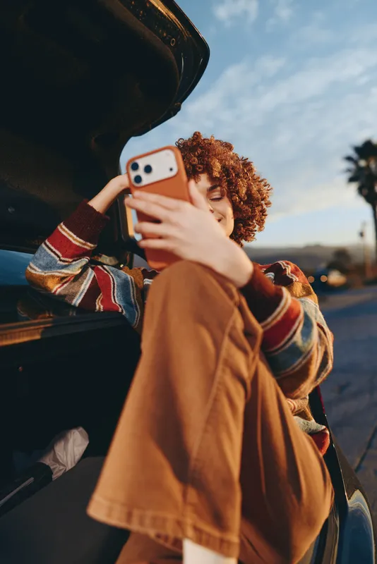 Woman lifestyle moment, curly hair, smartphone selfie as she stands outdoors near a car on a street, travel mood in warm sunset daylight, casual coat, hand holding phone.