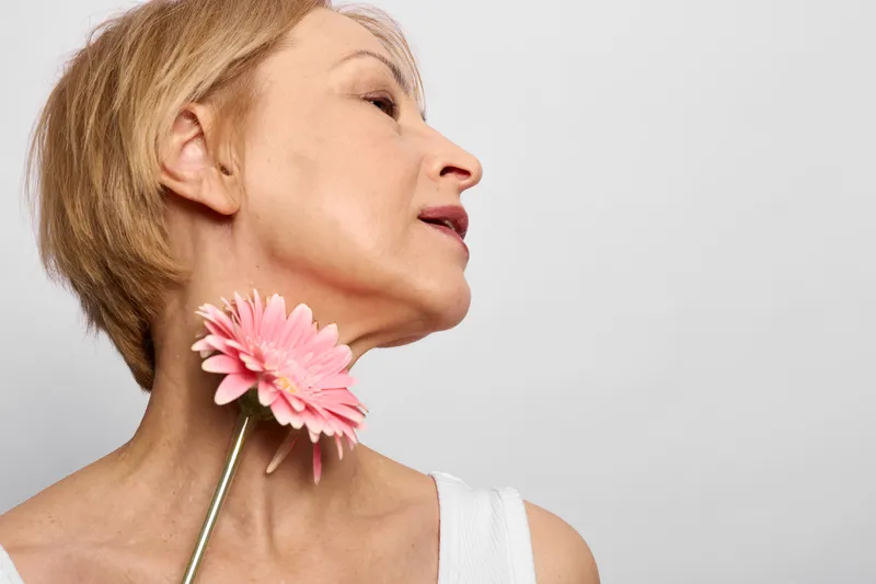 Woman closeup portrait, eyes closed, holding pink flower to her neck with stem, soft studio light, calm sensual beauty mood, smooth skin, minimal background