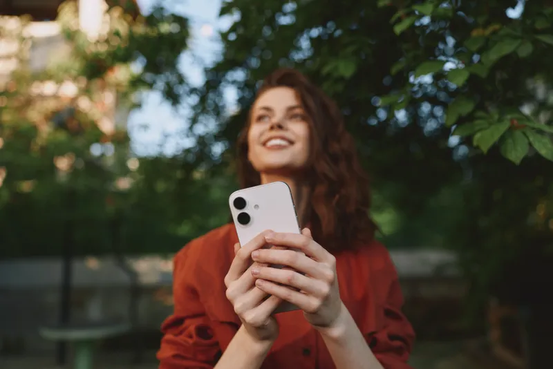 Young woman with curly hair holds a phone outdoors, smiling up at the sky amid lush greenery, capturing a candid lifestyle moment