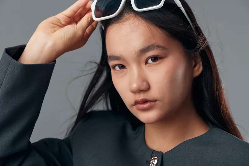 Young woman with long dark hair wearing black jacket and white sunglasses adjusting eyewear with hand, looking at camera with confident expression on neutral background.