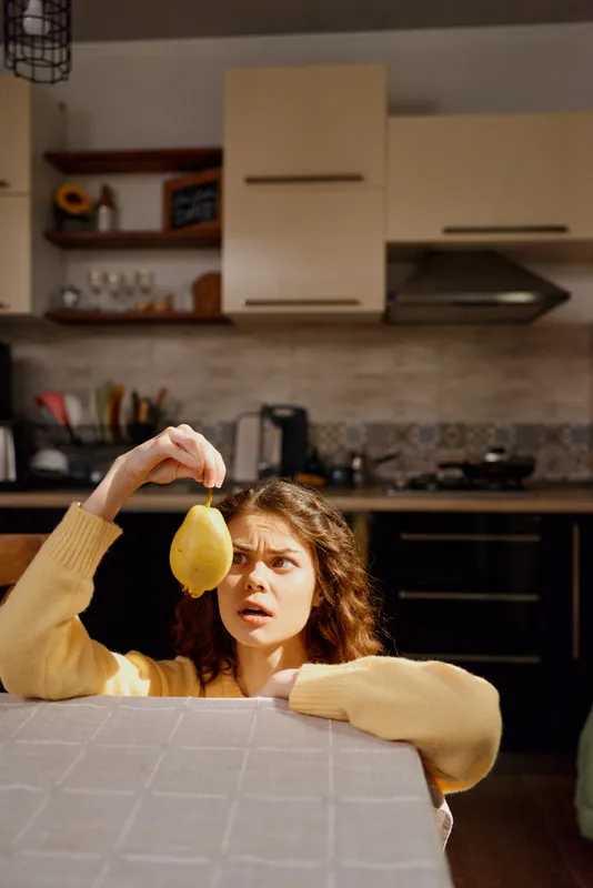 Smiling woman in a yellow sweater holds a lemon over a kitchen counter, surprised by its scent. Bright indoor lighting captures a warm home moment, a casual lifestyle scene in a cozy, domestic
