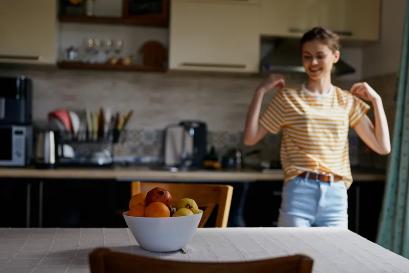 Cheerful woman in a cozy kitchen, casual striped top, smiling near a fruit bowl on a wooden table, warm natural light, relaxed home moment captured up close