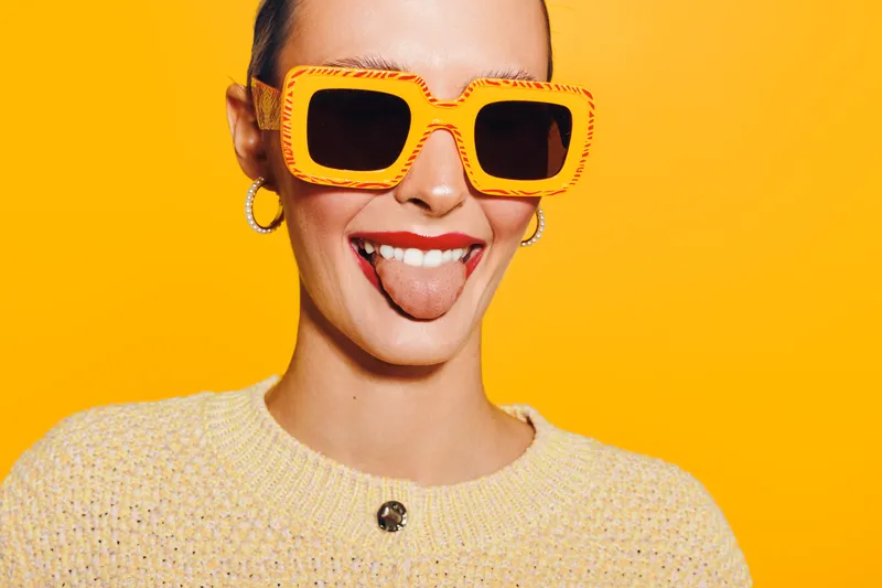 Young woman with orange sunglasses and yellow sweater sticks out tongue on bright yellow background. Playful mood, summer style, and fun expression in studio portrait.