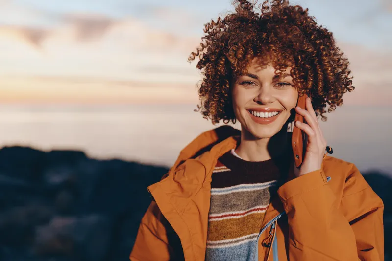 Woman lifestyle moment, smiling curly hair woman, phone to ear, wearing orange jacket, outdoors near sunset, mountains in soft blur, warm travel atmosphere, casual portrait