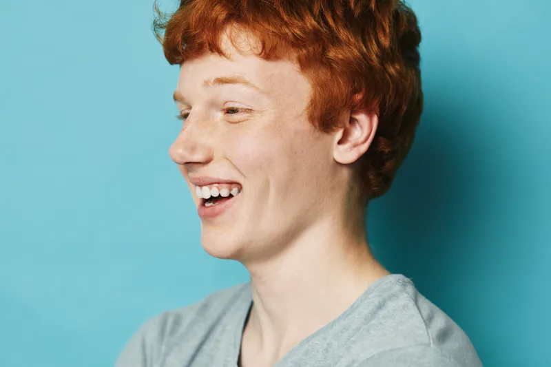 Young man with red hair smiling and laughing looking away against blue background in casual shirt, happy expression and natural portrait indoors.