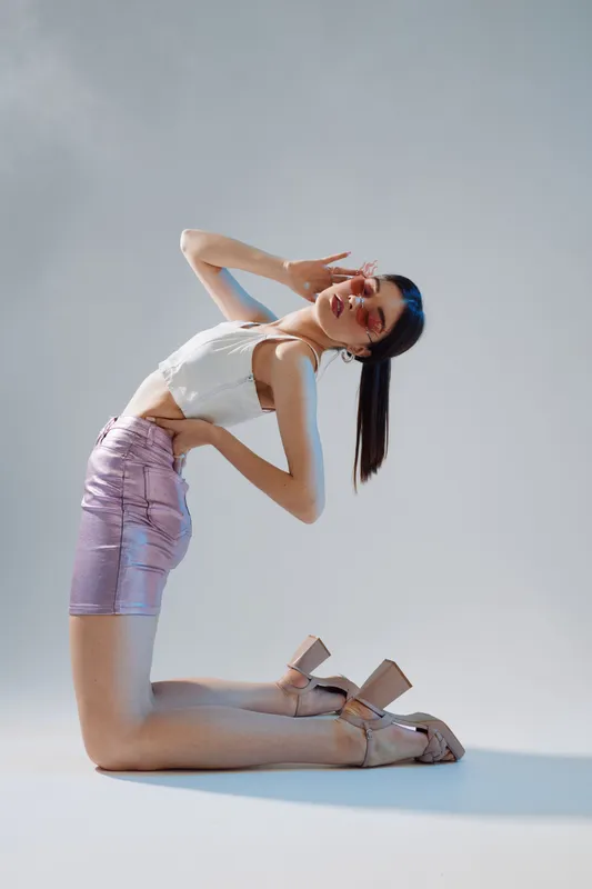 Young woman in a white top and shiny skirt poses kneeling on the floor against a light background. Concept of beauty, fashion, style, and modern lifestyle with confident expression.