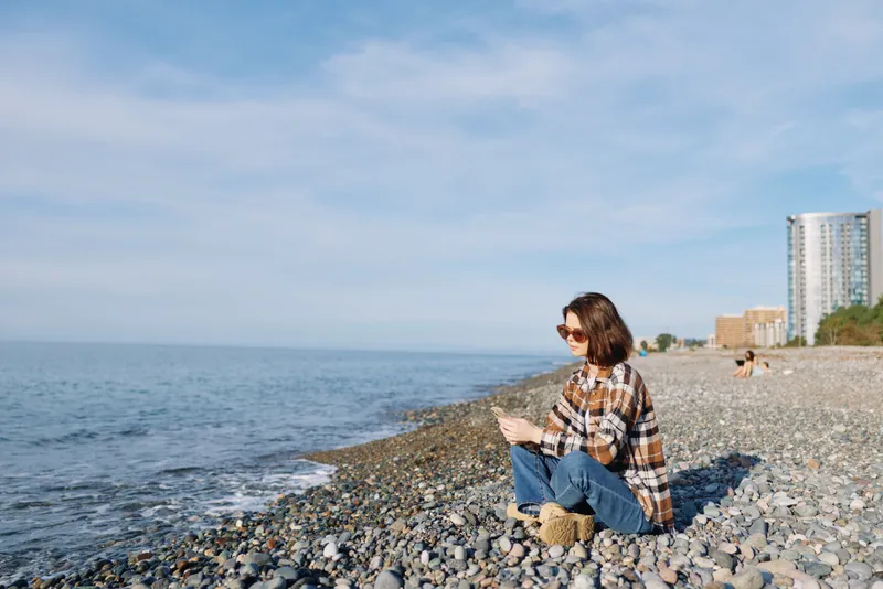A woman sits on a pebble beach by the sea, wearing a plaid shirt and sunglasses, relaxing and enjoying the coastal view with calm water and distant buildings.