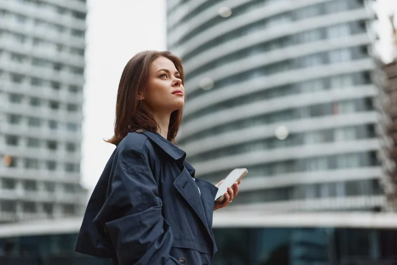 Young woman in a dark trench coat stands outdoors in an urban setting, looking up with a phone in hand, a confident candid moment.