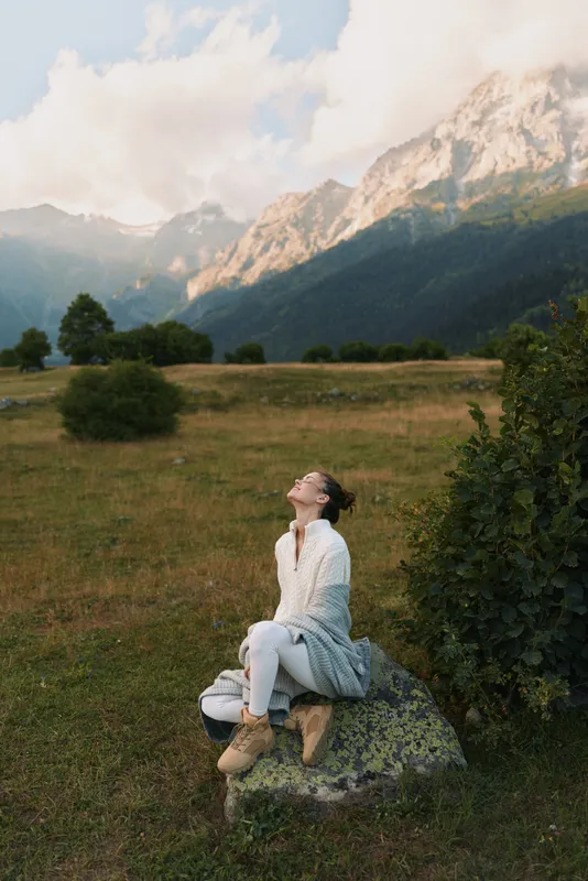 One young woman with brunette hair wears a white cardigan and sits on a rock in a meadow, gazing upward at distant mountains with a calm, reflective mood.