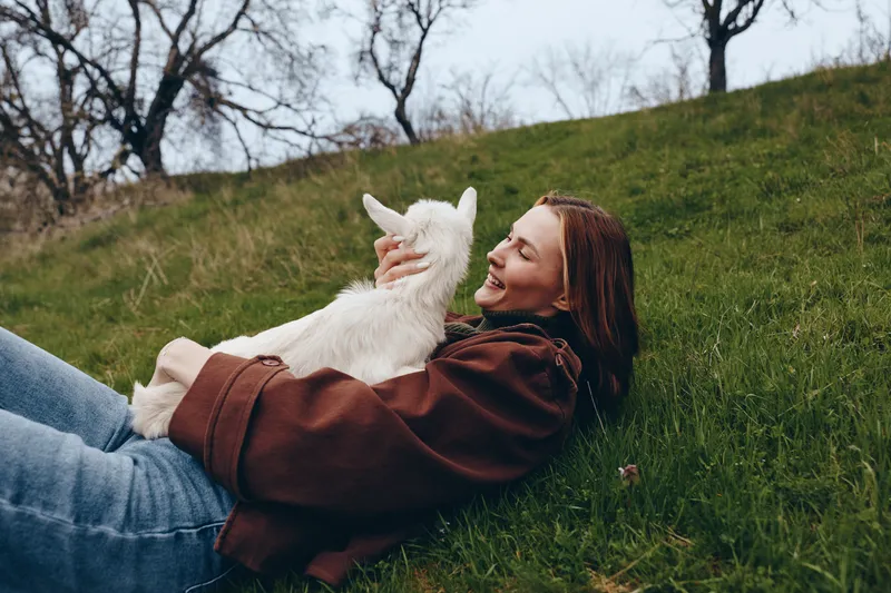 A young woman in a brown jacket lies on a grassy hill holding a white baby goat while smiling and enjoying a peaceful moment in a natural outdoor setting.