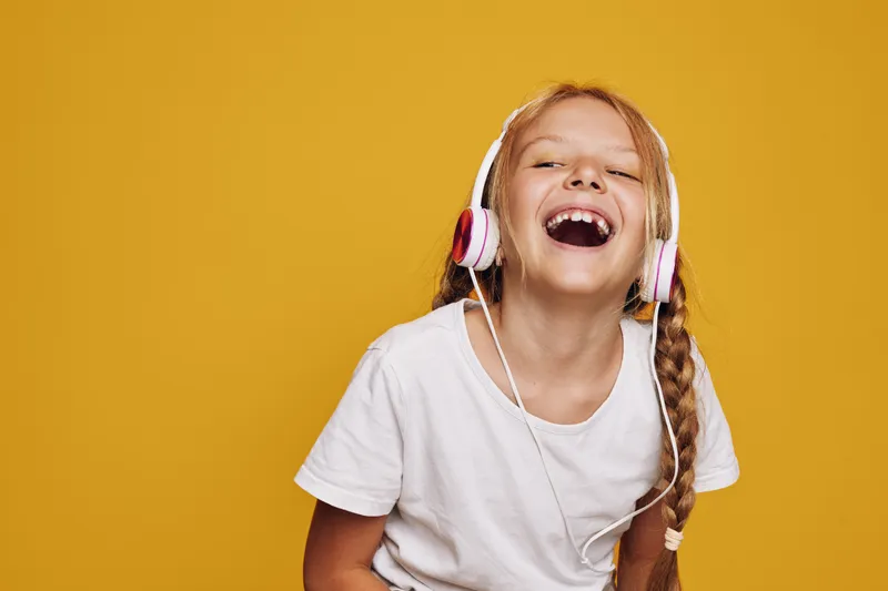Happy girl with braided hair wearing white headphones laughs joyfully against a yellow background. Studio portrait capturing fun, music, and carefree childhood moments.