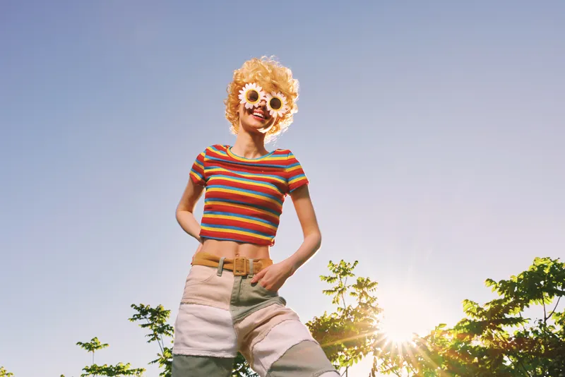 Smiling woman in a striped crop top and high waist shorts stands under a bright blue sky, retro film color mood, sunny outdoors, showcasing a carefree lifestyle and fashion.