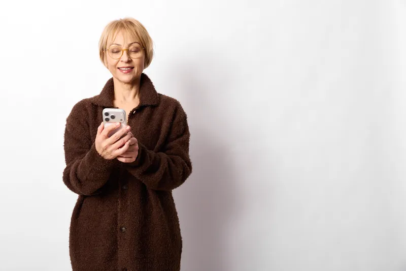 Smiling senior woman, selfie with smartphone, portrait in a white studio, casual fleece jacket, soft studio lighting, friendly self care mood, today smile