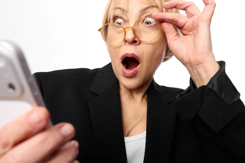 Woman surprised holding a smartphone and adjusting glasses, closeup older portrait with shock, alarmed expression on white background, tech communication in studio light