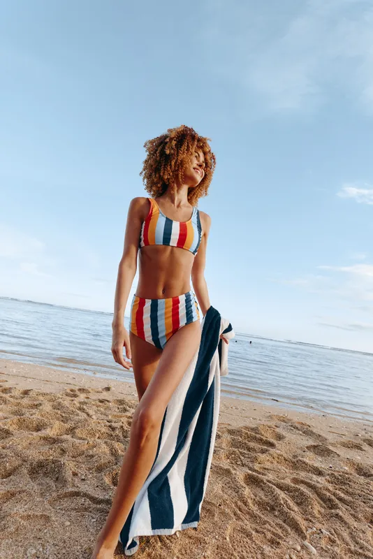 Beach, woman with curly hair in striped swimsuit walks along sand holding a folded towel, ocean horizon behind, bright summer light, calm waves, concept of relaxation and freedom in travel