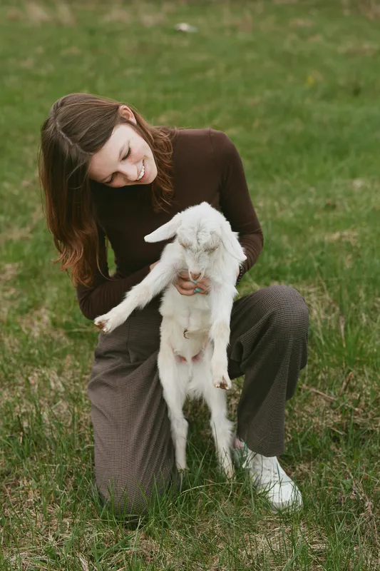 Bright outdoor moment with a woman kneeling in a meadow, cradling a young goat, warm sunlight, relaxed posture, casual clothing, natural colors, serene connection with nature