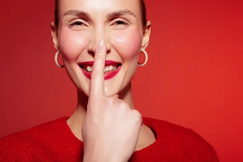 Smiling woman with red lipstick and hoop earrings touches her nose with finger against red background. Close-up portrait with bright lighting and cheerful expression. Concept of happiness and beauty.
