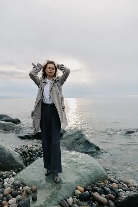 Woman in long coat stands on rocky shore with hands in hair, looking confidently ahead. Calm sea and cloudy sky background create peaceful and contemplative mood.