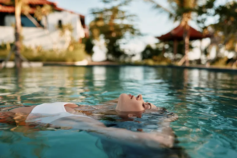 Young woman with long hair floats relaxed on her back in a clear swimming pool wearing a white swimsuit during a sunny day in a tropical lifestyle setting.