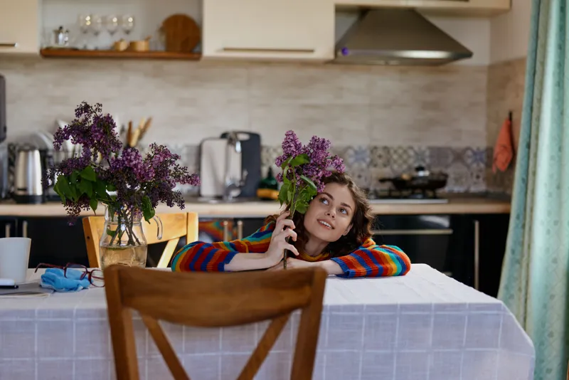 Smiling woman leaning on a kitchen table with a vase of lilac flowers. Cozy home scene, casual lifestyle, everyday moment, warm mood, natural light, relaxed composition, intimate mood.