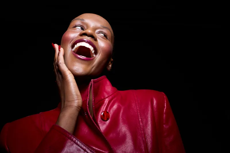 Happy African woman with dark skin wearing red leather jacket smiles joyfully with hand on face. Black background highlights her radiant expression and confident mood in studio portrait.
