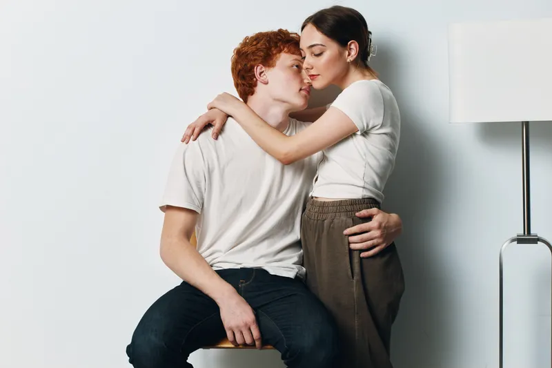 Couple embracing in a tender moment with eyes closed, woman standing and man sitting on stool in casual clothing in interior with white wall and lamp.