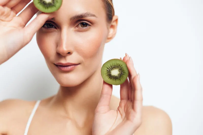 Young woman with clear skin holds two halves of a kiwi fruit near her face while looking confidently at the camera in a bright studio setting.