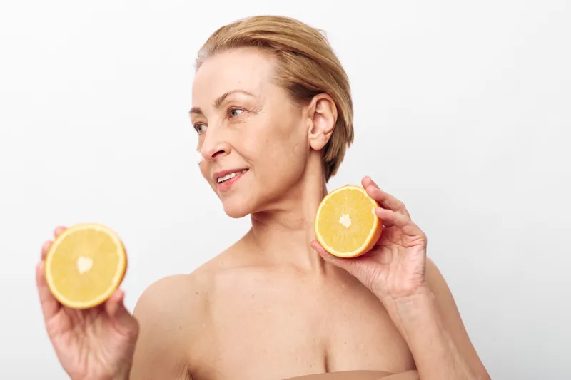 Mature woman with short hair holds two halves of fresh orange fruit near face, smiling softly on white background. Concept of healthy lifestyle, natural beauty, skincare, and wellness.