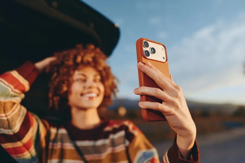 woman lifestyle selfie smartphone outdoors, curly hair smile, holding orange phone for a selfie, sky and sunset horizon behind, casual street lifestyle moment in golden daylight