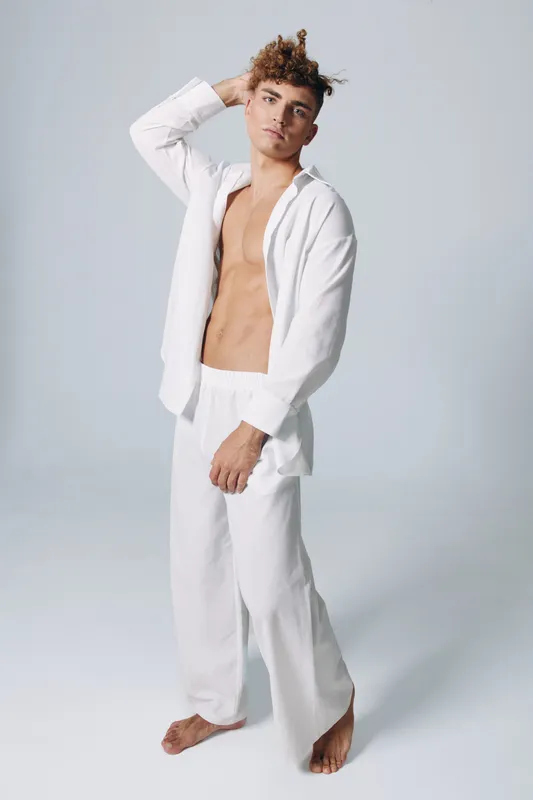 Young man with curly hair posing barefoot in white open shirt and loose pants against plain background, casual fashion and relaxed style with confident expression.