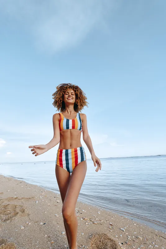 Happy woman walking on a sandy beach wearing a colorful striped swimsuit, ocean waves behind her, bright summer sunlight in a clear blue sky, outdoors lifestyle and freedom concept