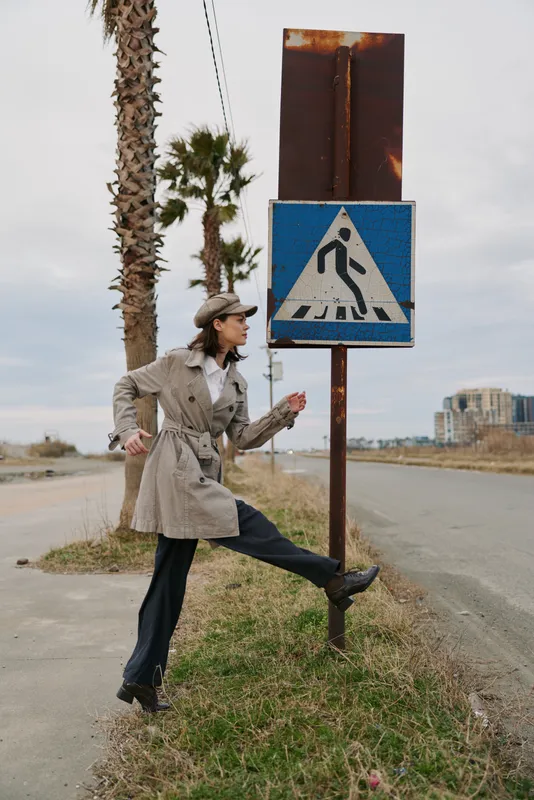 Woman in trench coat and cap poses playfully near pedestrian crossing sign on roadside with palm trees and urban buildings in background. Concept of fashion, street style, and urban life.