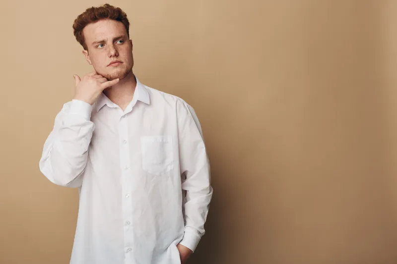 Thoughtful young man in white shirt looks away with hand on chin against beige background. Concept of contemplation, decision making, and introspection in casual style.
