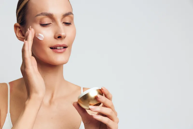 Young woman with clear skin applies cream on face holding open jar isolated on white background skincare beauty routine moisturizing treatment concept.