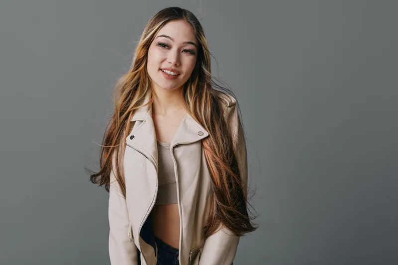 Young woman with long hair wearing light jacket smiling softly and looking to side on gray background in studio portrait with natural expression.