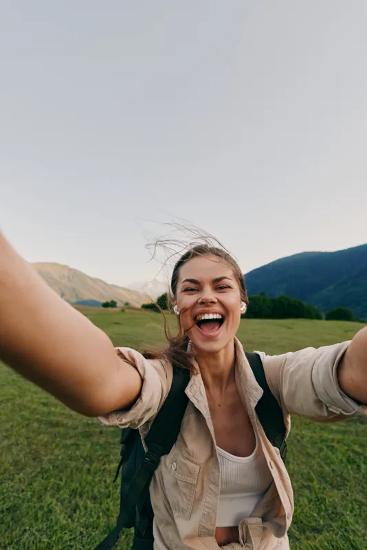 Vibrant woman outdoors, smiling with arms open, backpack ready, sunlit field and mountains behind, carefree lifestyle, candid moment, natural colors