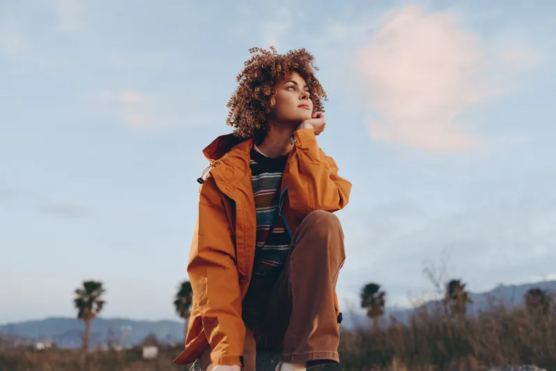 Woman lifestyle outdoors, orange jacket, curly hair, standing in open field at sunset evening, thoughtful look, natural daylight, blurred landscape background, casual portrait