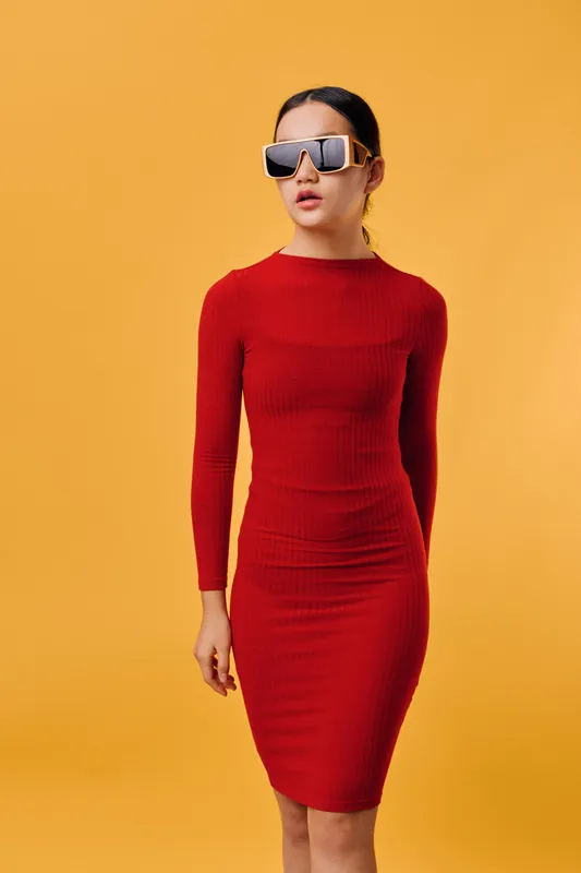 Woman wearing tight red dress and large sunglasses standing against yellow background. Fashion model with serious expression and stylish outfit for modern look and style.
