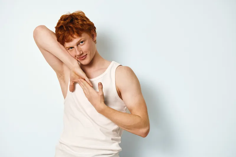 Redhead young man in sleeveless white shirt posing with playful expression touching arm against a light background. Casual style, smiling and confident portrait in studio setting.