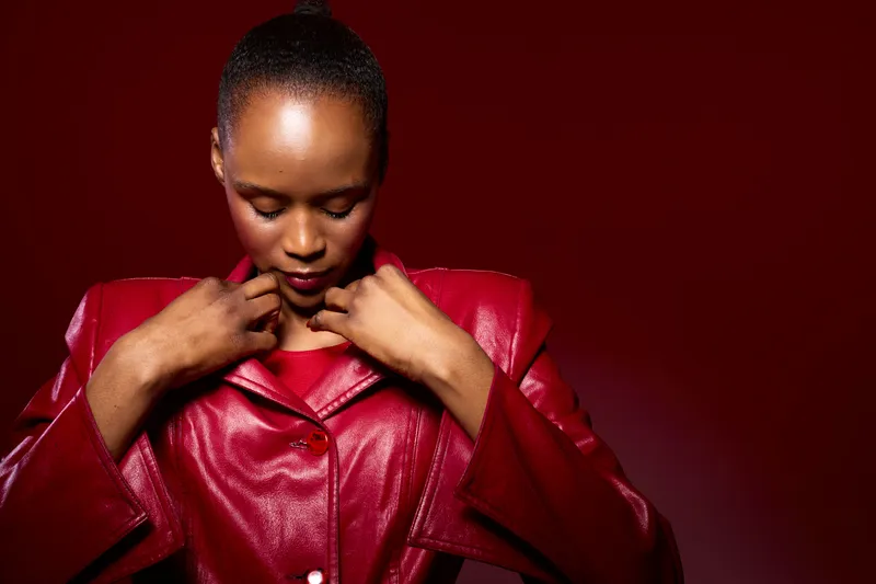 Portrait of a young African woman with dark skin wearing a red leather jacket adjusting collar. Studio shot on red background. Concept of fashion, confidence, and style for dark-skinned models.
