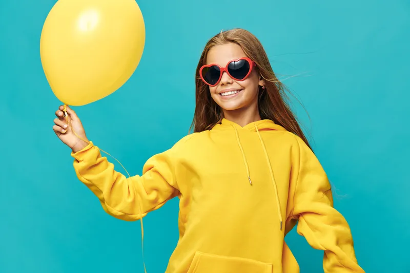 Girl in yellow hoodie holding yellow balloon smiling wearing heart shaped sunglasses on blue background with long hair and casual style for celebration and happiness.