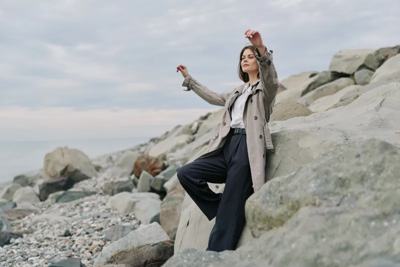 Young woman in stylish coat and wide pants stands on rocky beach with cloudy sky. Relaxed pose and nature background create mood of calm and contemplation outdoors.