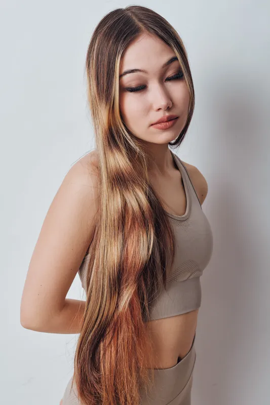 Young woman with long hair standing in studio against white wall wearing beige outfit looking down with calm expression and natural makeup.